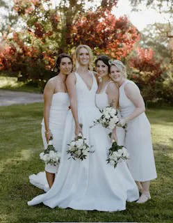 Alex and two bridesmaids in white dresses holding white bouquets beside an autumn tree.
