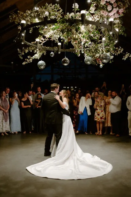 Bride and groom dancing beneath a suspended floral ring with greenery and hanging glass ornaments.