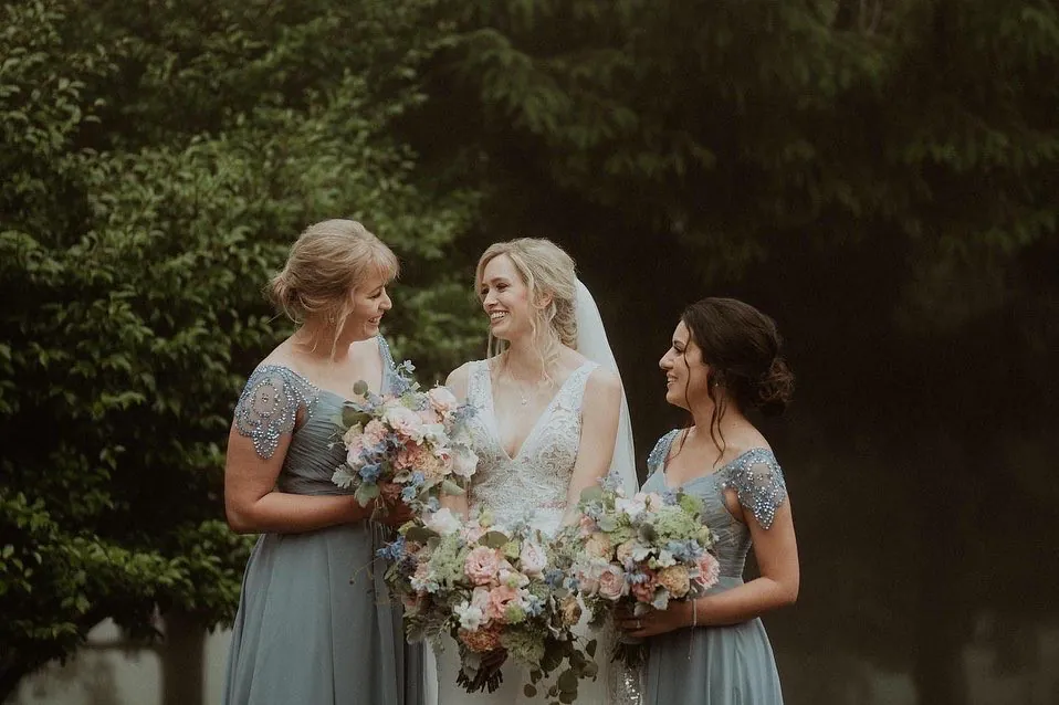 Bride and two bridesmaids in pale blue dresses holding pastel bouquets in a garden.