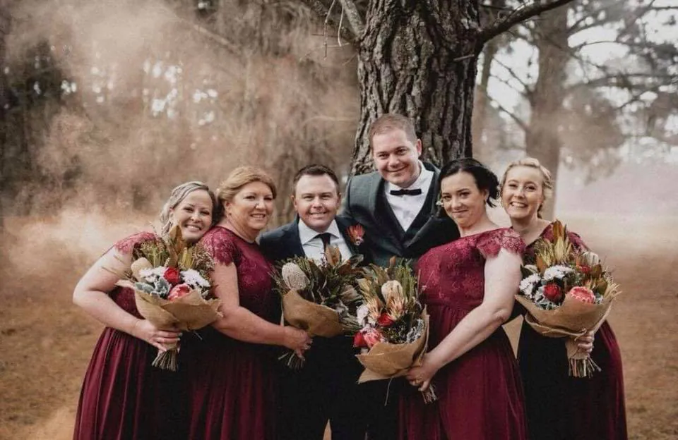 Bride and groom with their wedding party in burgundy dresses holding mixed floral bouquets beneath a tree.