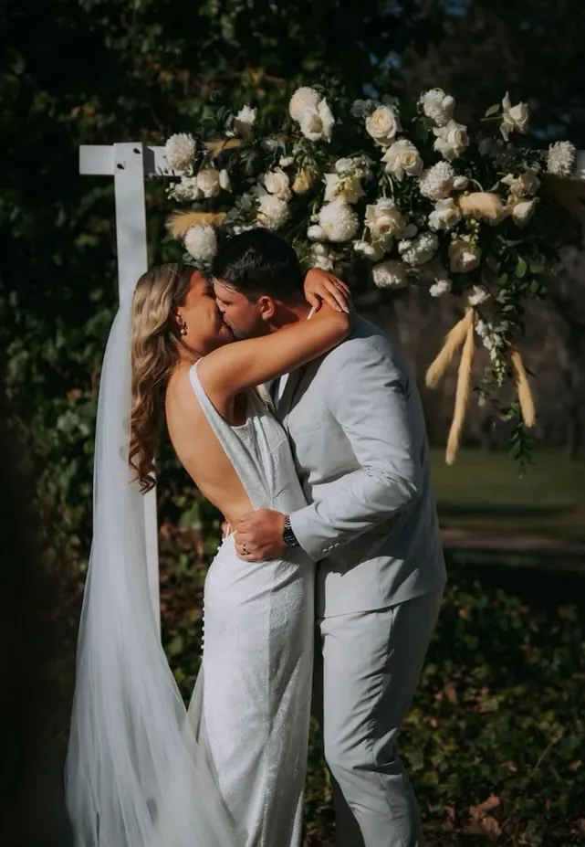 Gemma and her groom kissing beneath a white arbour with cream and white flowers in a garden.