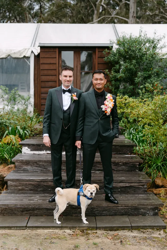 James and David in dark suits with floral lapel arrangements and their dog on timber steps.