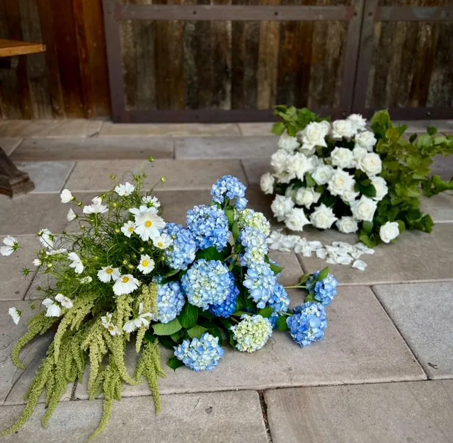 Blue hydrangeas, white flowers and trailing greenery arranged at ground level for a wedding ceremony.