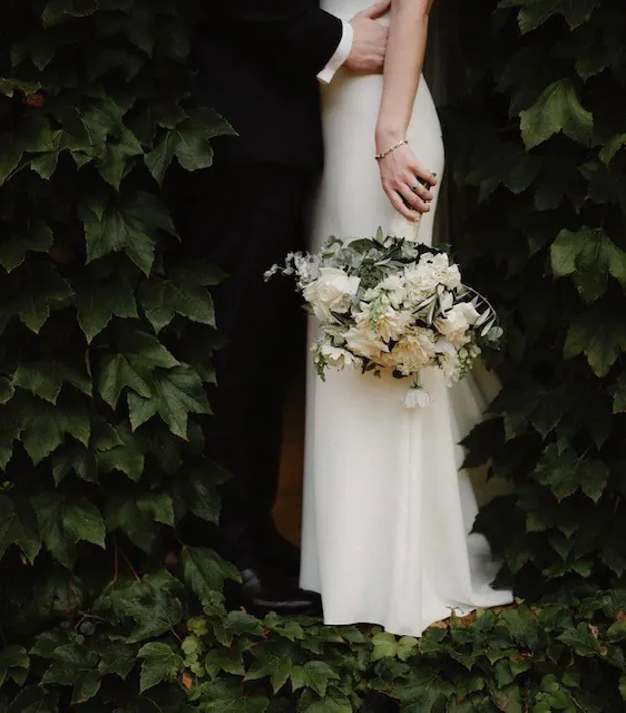 Close view of Olivia holding a white bouquet with long ribbons beside green ivy.