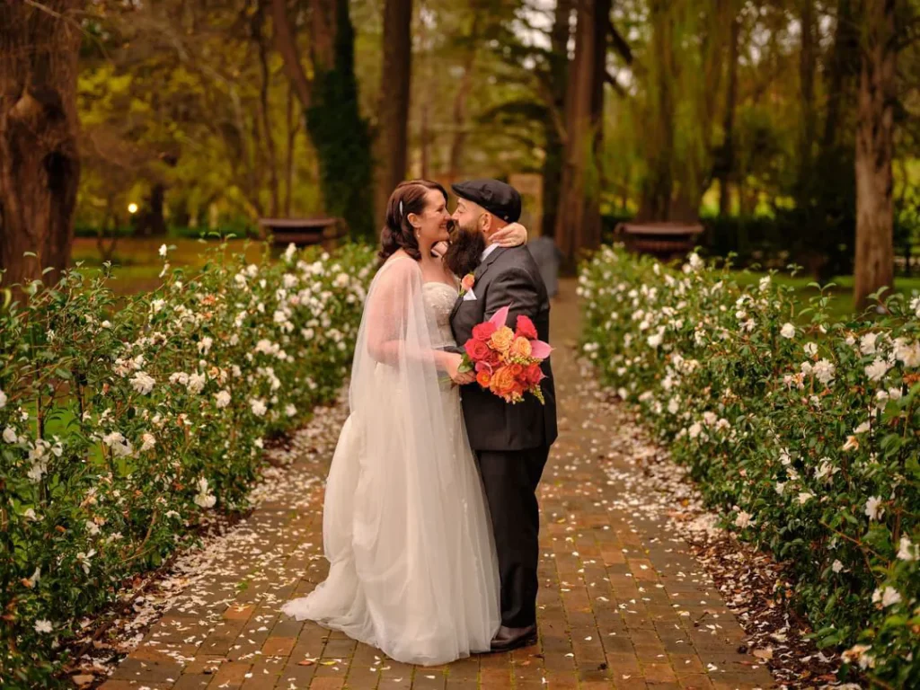 Sarah and Tony kissing on a tree lined path with a vivid orange, pink and red bridal bouquet.