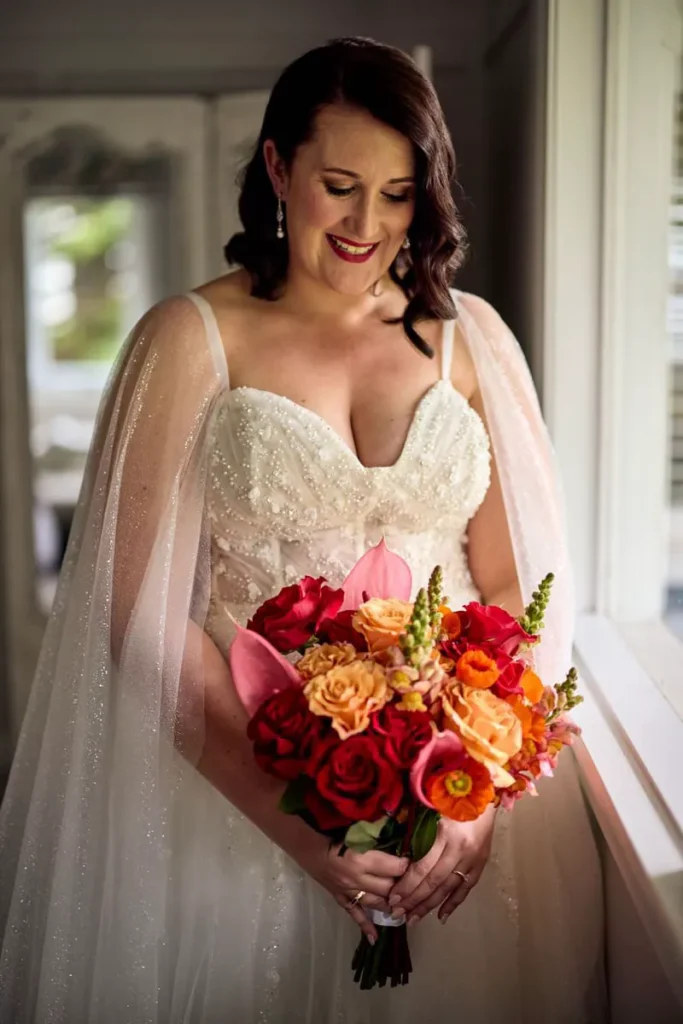 Sarah holding a bright bouquet of orange, pink, yellow and red flowers beside a window.