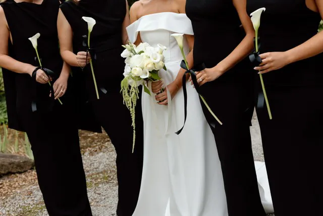 Bridesmaids in black dresses holding single white calla lilies beside a bride with a white bouquet.