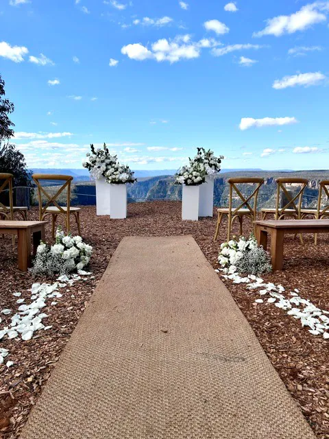 White flowers on two white plinths framing an outdoor ceremony aisle beneath a blue sky.