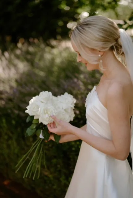 Bride looking down at a white rose bouquet with long green stems in a garden.